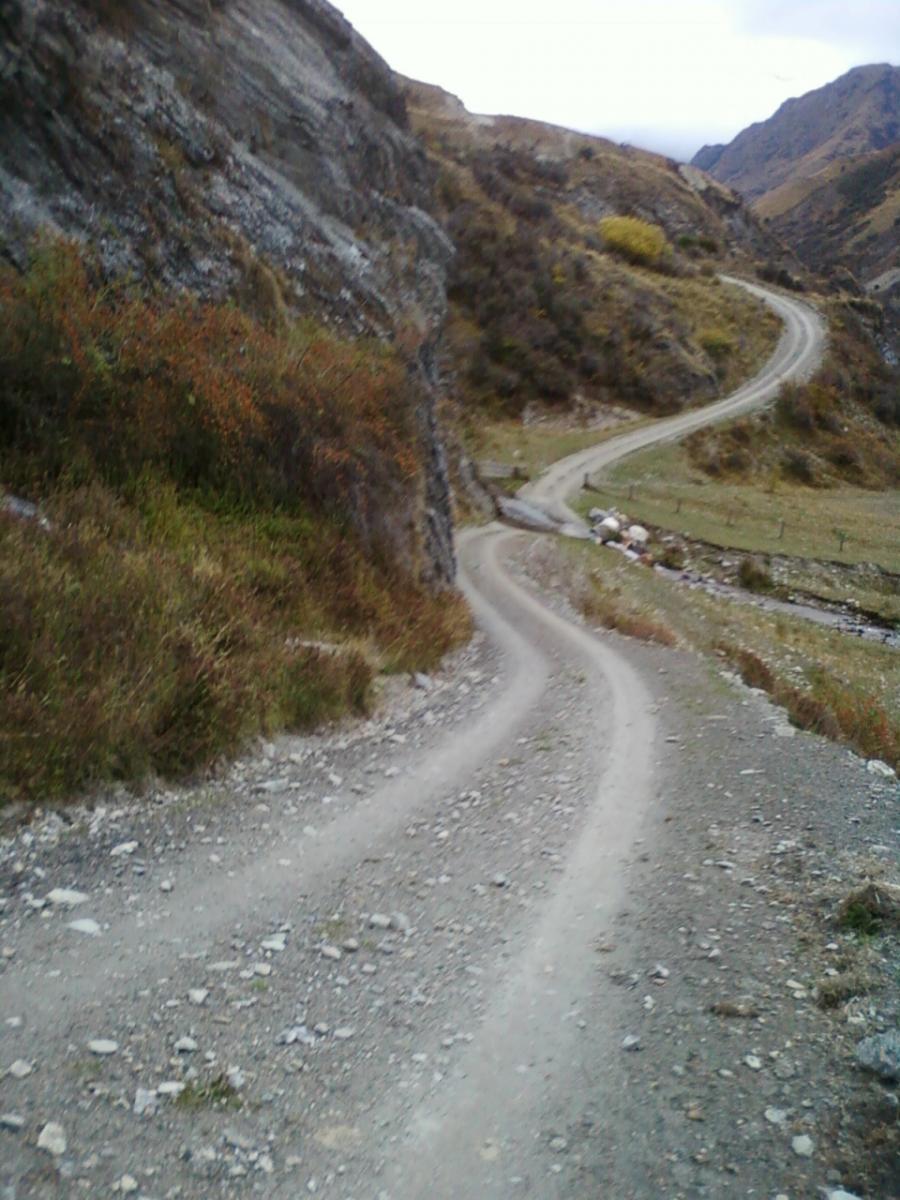 A winding dirt road cutting through a rugged landscape, flanked by rocky terrain and sparse vegetation. The path leads into the distance, surrounded by hills and mountains under a cloudy sky. Moonlight Circuit mountain bike trail.