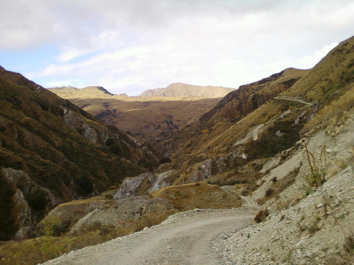 A winding dirt road cuts through a rugged valley, flanked by steep hills and rocky terrain. The landscape features a mix of grassy slopes and rocky outcrops under a partly cloudy sky, with distant mountains visible in the background. Moonlight Circuit mountain bike trail.