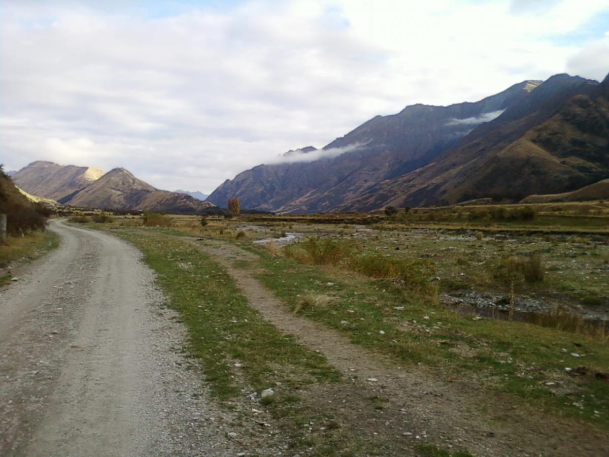 A winding gravel road surrounded by mountains under a partly cloudy sky, with grassy areas and a small stream visible along the side. Moonlight Circuit mountain bike trail.