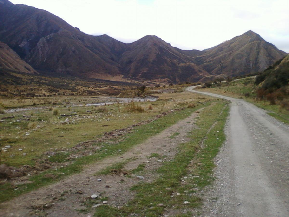 A winding gravel road leads through a rural landscape, flanked by grassy areas and patches of stones. In the background, a range of rugged mountains rises under a cloudy sky, highlighting the natural beauty of the scene. Moonlight Circuit mountain bike trail.