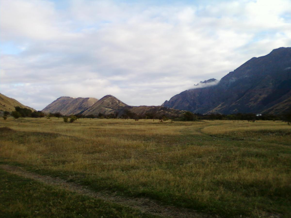 A scenic view of rolling hills and mountains under a cloudy sky, with tall grasses and sparse trees in the foreground. The landscape features varying elevations, creating a natural and tranquil setting. Moonlight Circuit mountain bike trail.