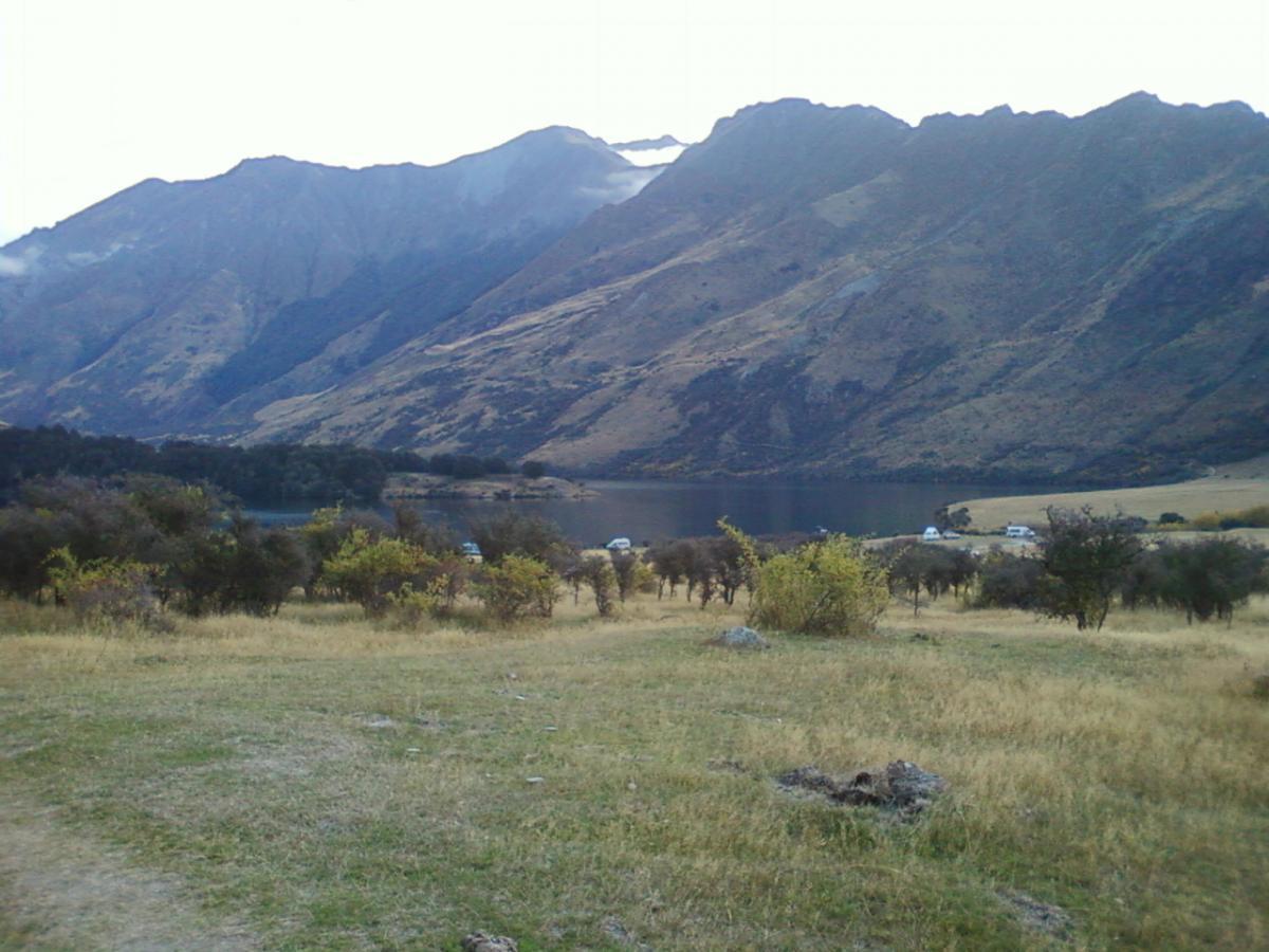 A scenic view of mountains and a lake, with grassy terrain and sparse trees in the foreground. The landscape features rolling hills and peaks, with some clouds hovering near the mountain tops. Moonlight Circuit mountain bike trail.