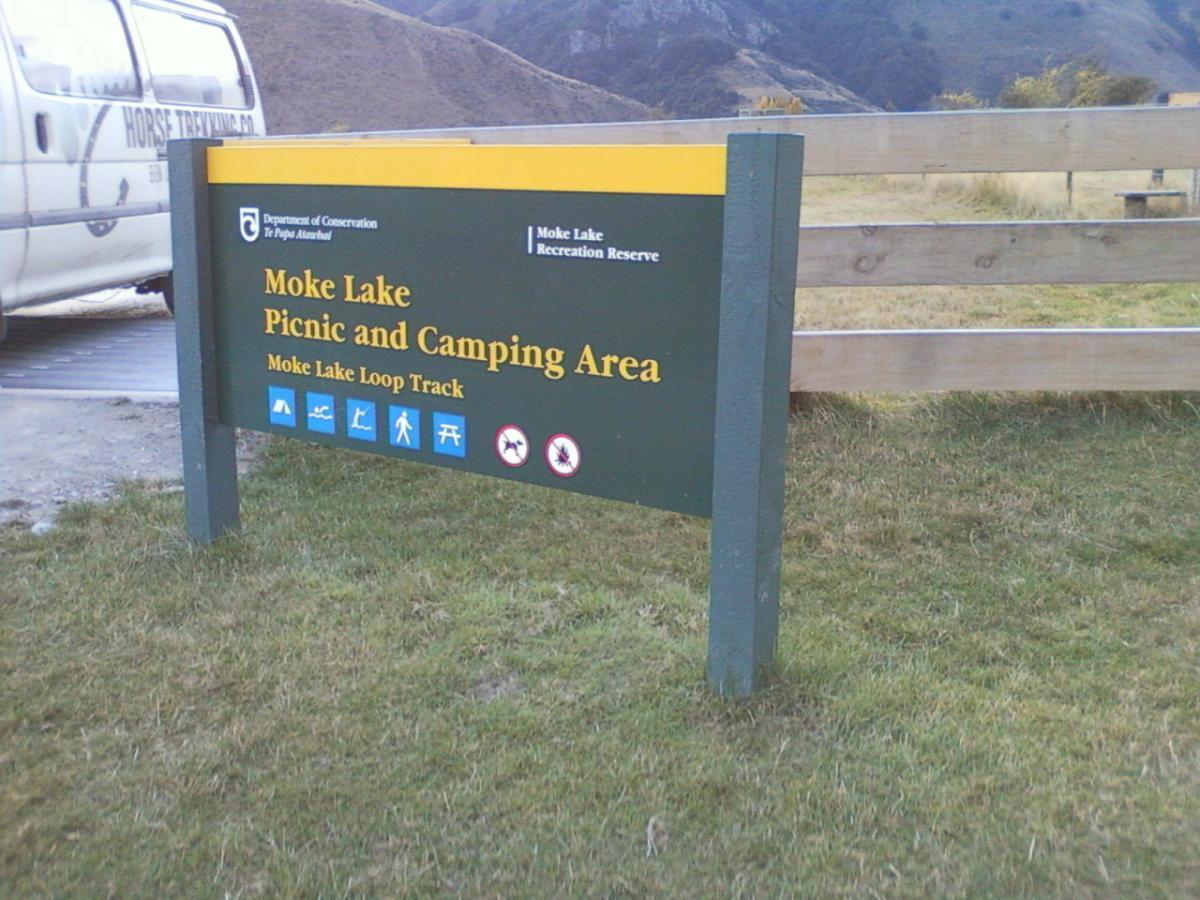 Sign for Moke Lake Picnic and Camping Area, featuring details about the recreational reserve and Moke Lake Loop Track, with icons indicating available activities. In the background, a parked vehicle can be seen along a gravel area, surrounded by grassy fields and hills. Moonlight Circuit mountain bike trail.