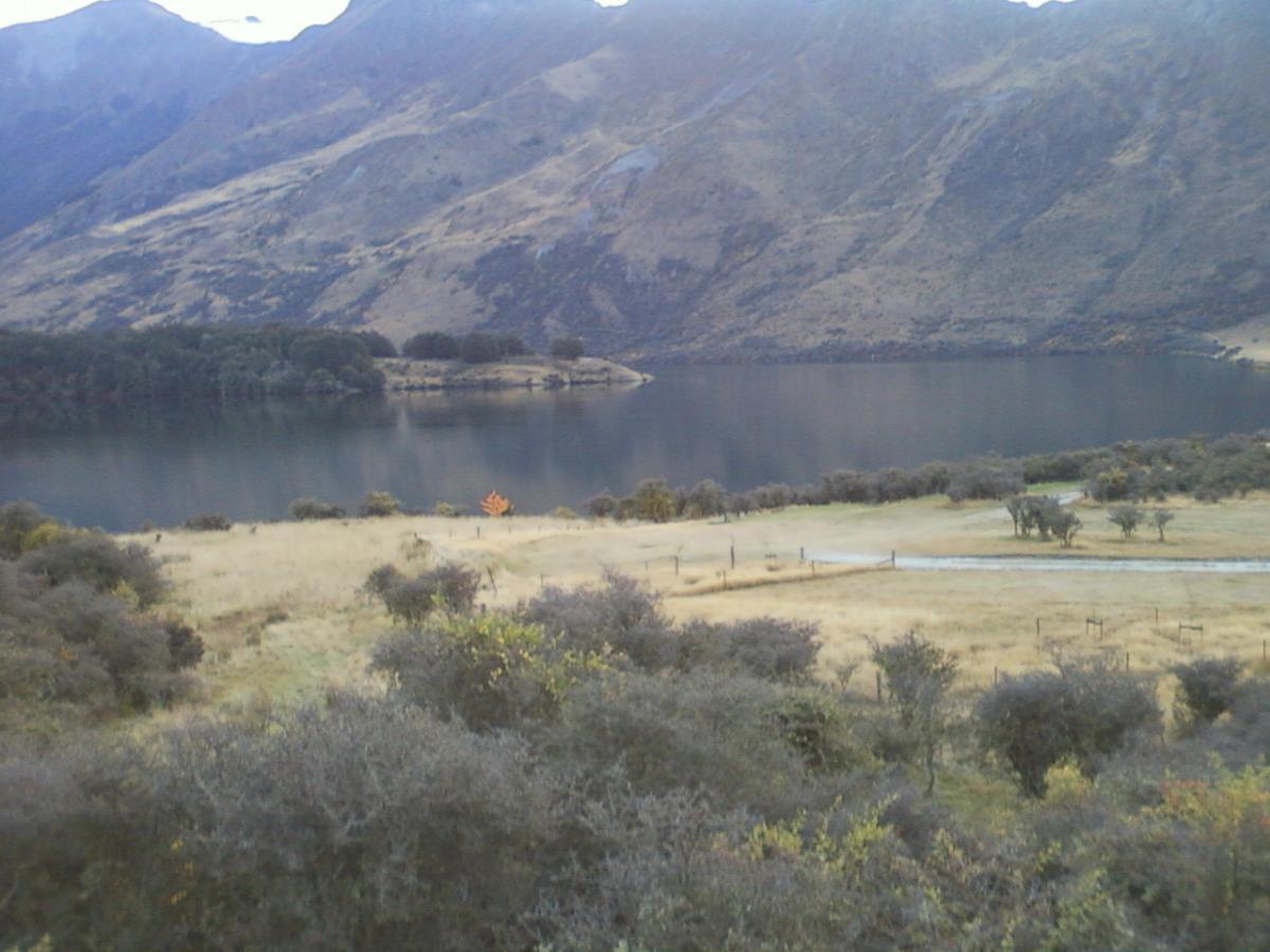 A tranquil landscape featuring a calm lake surrounded by gently sloping hills and mountains. In the foreground, a grassy area with sparse vegetation leads to the water's edge. The sky is overcast, and the scene captures a serene autumn atmosphere, highlighted by a single orange tree amidst the greenery. Moonlight Circuit mountain bike trail.