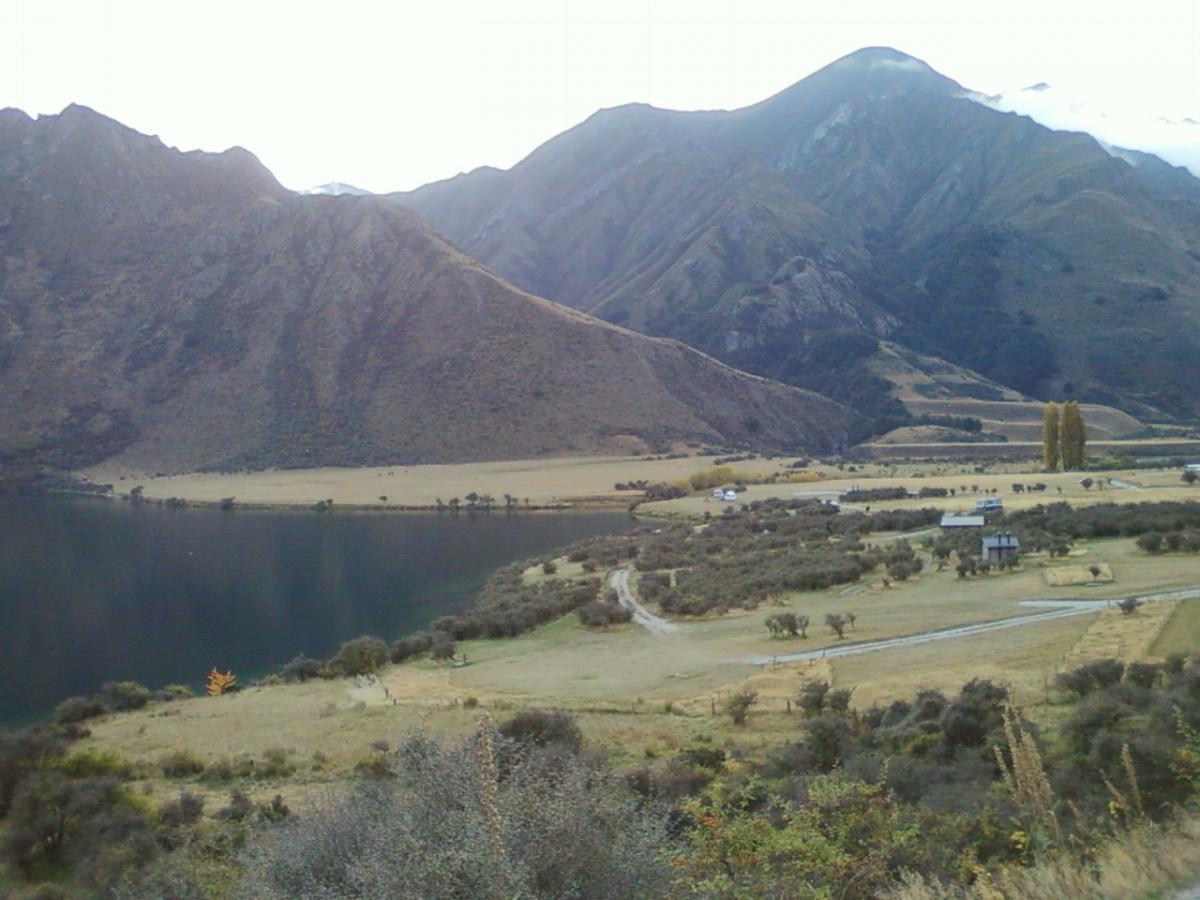 A scenic landscape featuring a tranquil lake surrounded by rolling hills and majestic mountains. The foreground displays a grassy area with scattered trees, while the background showcases steep, rugged mountain peaks partially shrouded in clouds. Small structures can be seen along the shoreline and in the valley, adding a touch of human presence to the serene natural setting. Moonlight Circuit mountain bike trail.