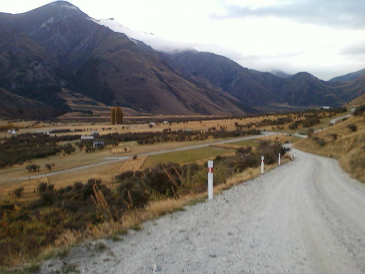 A gravel road winding through a mountainous landscape, surrounded by rolling hills and fields. Soft clouds hover over the peaks in the distance, while patches of greenery and autumn-colored trees dot the land below. Moonlight Circuit mountain bike trail.