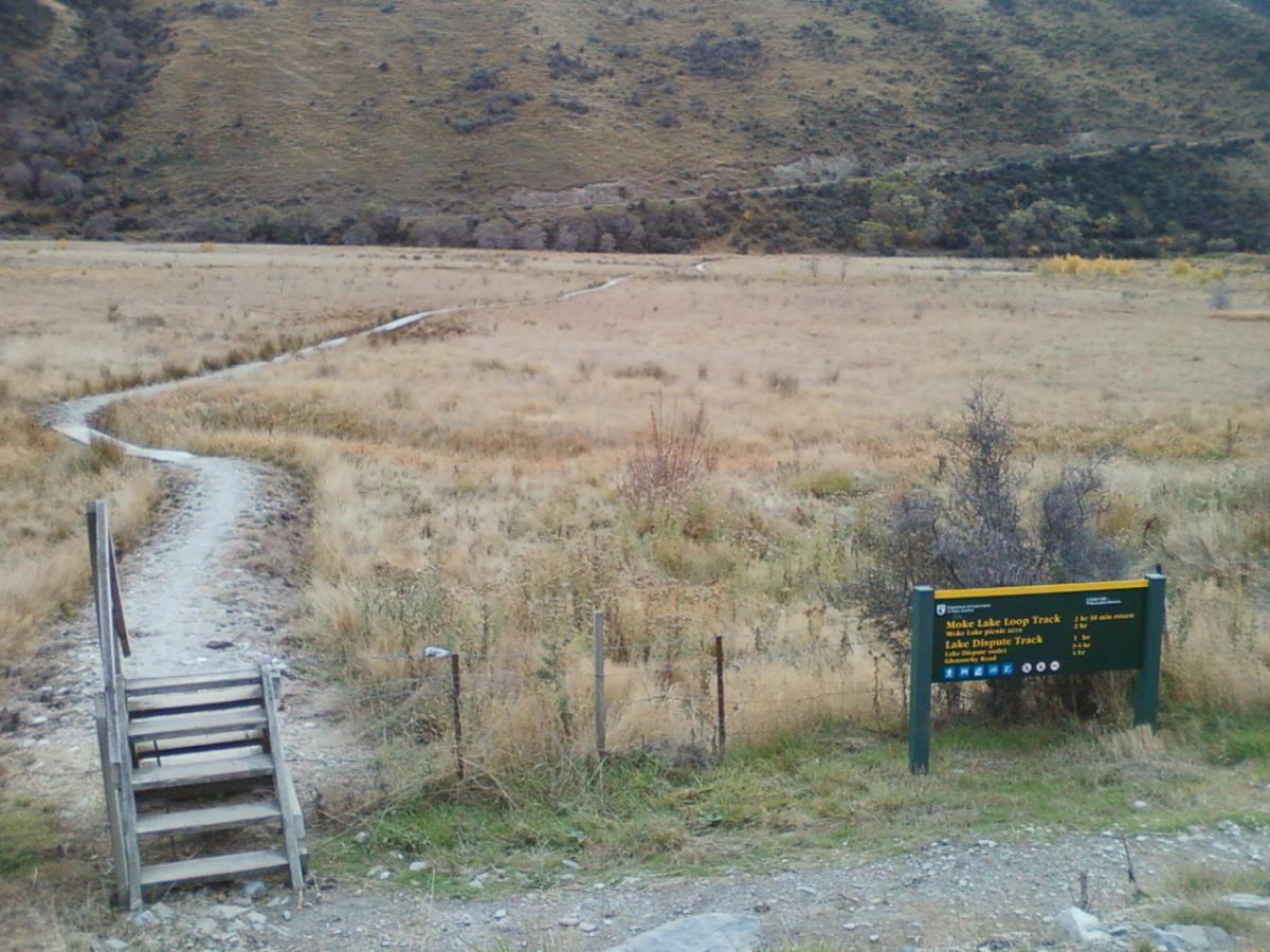 A winding dirt path leads through dry grassland towards distant hills, with a sign for the Moke Lake Loop Track and Lake Dispute Track prominently displayed in the foreground. The landscape features a mix of tall grass and sparse shrubs under a cloudy sky. A set of wooden steps is visible on the left, leading to the path. Moonlight Circuit mountain bike trail.