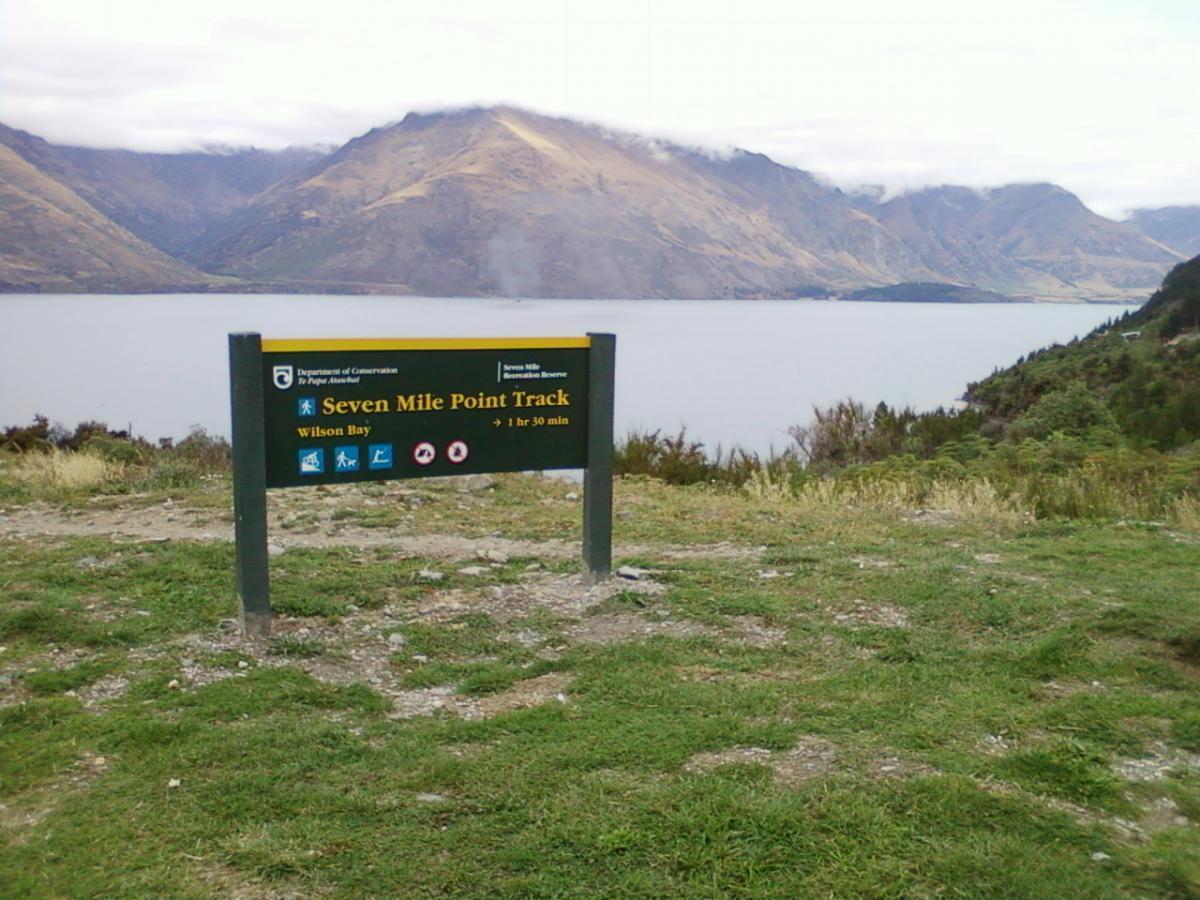 Sign for the Seven Mile Point Track located near Wilson Bay, surrounded by grass and mountains in the background, with a lake visible. The sign indicates a walking time of 1 hour and 30 minutes. Seven Mile Bike Park mountain bike trail.