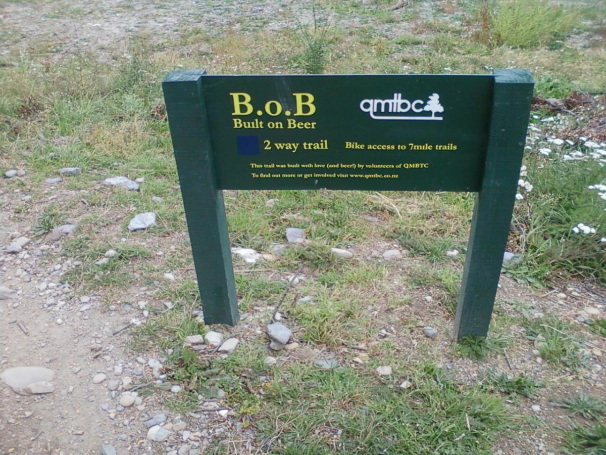 A sign indicating the "B.O.B. (Built on Beer)" trail, featuring a two-way bike access trail to 7-mile trails. The sign is dark green with white and blue text, displaying information about the trail's origin and encouragement to get involved with the QMBTC (Queenstown Mountain Bike Club). Surrounding grass and gravel are visible. Seven Mile Bike Park mountain bike trail.