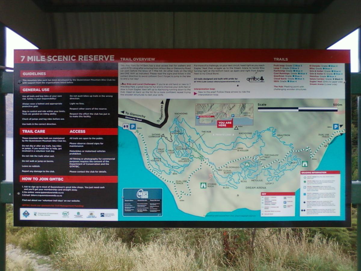 Alt text: A trail overview sign for the 7 Mile Scenic Reserve, featuring guidelines, trail care, access information, and a detailed trail map. The map includes designated biking trails, scale, and locations within the reserve, surrounded by grass and a shelter structure. 7 Mile Riding Area mountain bike trail.