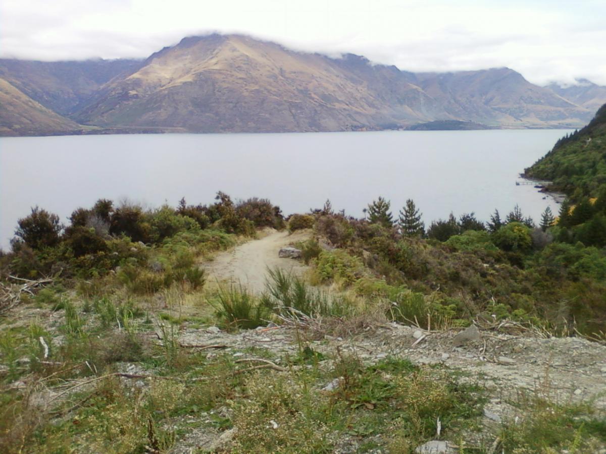 Scenic view of a tranquil lake surrounded by mountains and lush greenery, with a winding dirt path leading through the foreground. Soft clouds hover above the mountains in the distance. 7 Mile Riding Area mountain bike trail.
