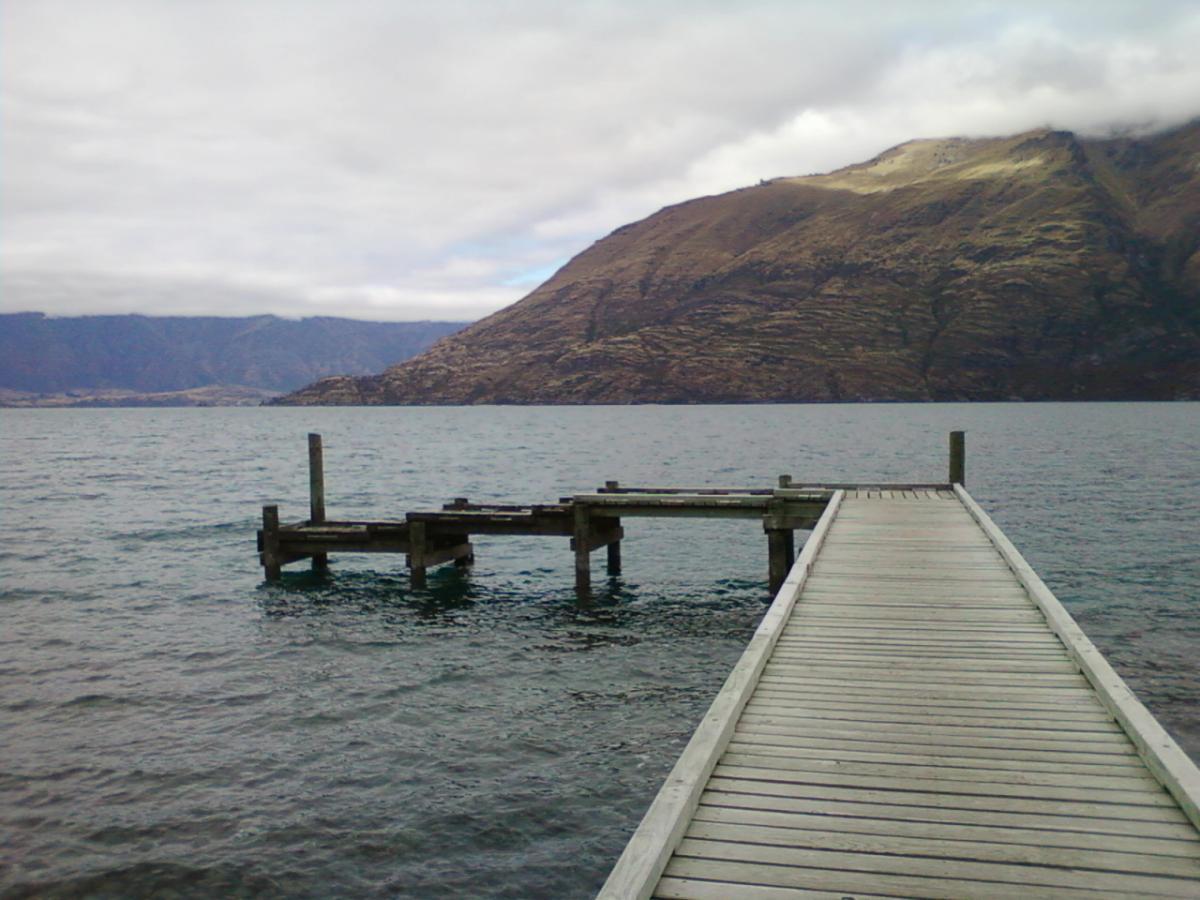 A wooden pier extends over calm water, leading toward a mountainous shoreline under a mostly cloudy sky. Gentle ripples in the water reflect the muted colors of the landscape. 7 Mile Riding Area mountain bike trail.