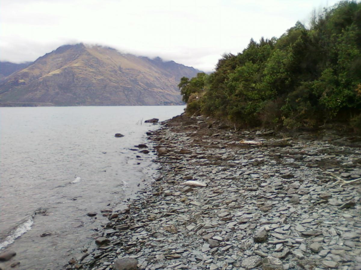 A rocky shoreline along a calm lake, with low waves lapping at the stones. In the background, a partially cloud-covered mountain rises above the water, surrounded by lush greenery. The sky is overcast, creating a serene and tranquil atmosphere. 7 Mile Riding Area mountain bike trail.