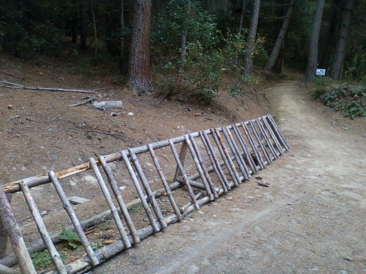 A wooden barrier made of logs is placed along a dirt path in a forested area, with trees and underbrush visible in the background. A small sign can be seen in the distance, indicating the direction of the trail. 7 Mile Riding Area mountain bike trail.