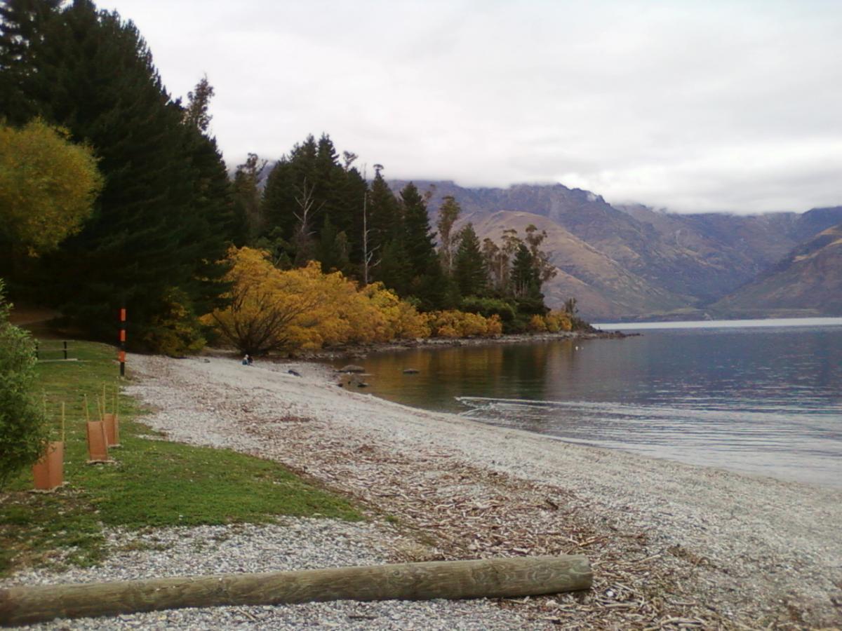 A tranquil lakeside scene featuring a pebbly beach bordered by lush green and autumn-colored trees. The calm water reflects the surrounding mountains, which are partially shrouded in clouds. A few pieces of driftwood are scattered along the shore, and a person can be seen sitting by the water's edge, enjoying the serene landscape. 7 Mile Riding Area mountain bike trail.
