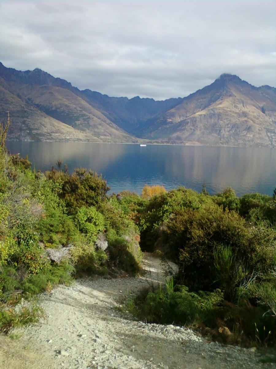 A scenic view of a lake surrounded by mountains, with a winding gravel path leading down through lush green foliage in the foreground. The sky is partially cloudy, reflecting soft light on the water's surface. 7 Mile Riding Area mountain bike trail.