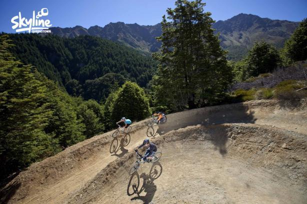 Three mountain bikers navigate a dirt track surrounded by lush trees and steep mountains under a clear blue sky. The scene captures the thrill of outdoor biking adventures in a scenic landscape. Queenstown Bike Park mountain bike trail.