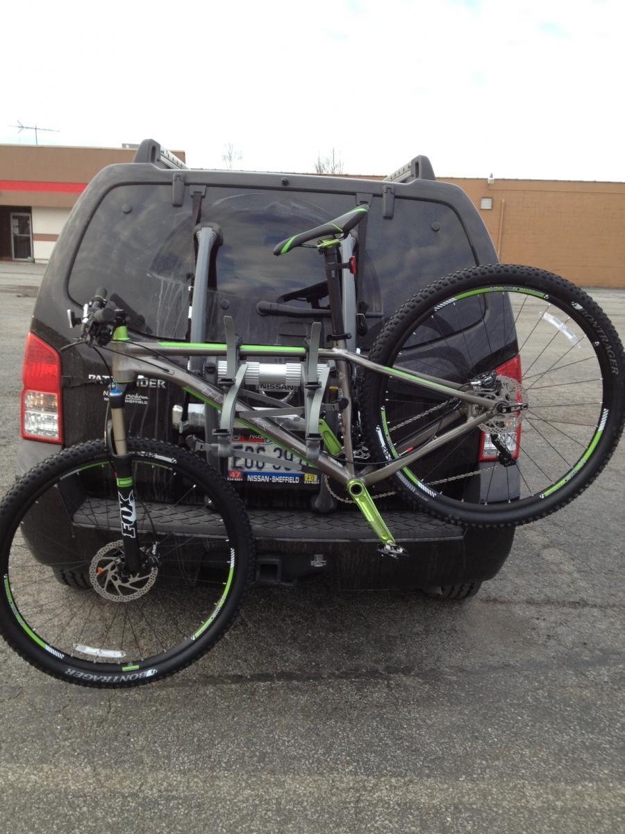 Trek Stache 8: A mountain bike is securely mounted on the back of a black SUV, with part of the vehicle's rear visible. The bike features distinctive green accents and is attached to a bike rack, set against an empty parking lot backdrop on a cloudy day.