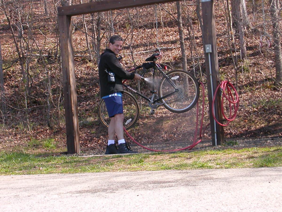 A person is using a hose to wash a bicycle that is suspended on a bike rack in a wooded area. The individual is smiling and wearing a black jacket, shorts, and athletic shoes. Leaves cover the ground, and the background features trees with no leaves, indicating early spring or late fall. Angler's Ridge mountain bike trail.