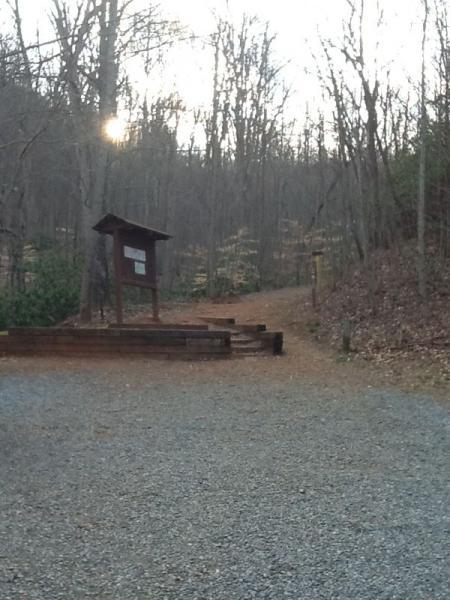 A gravel path leading into a wooded area, with bare trees and a signboard on the left. The sun is faintly visible in the background, indicating early morning or late afternoon light. The scene conveys a tranquil outdoor setting. Aska Trail System mountain bike trail.