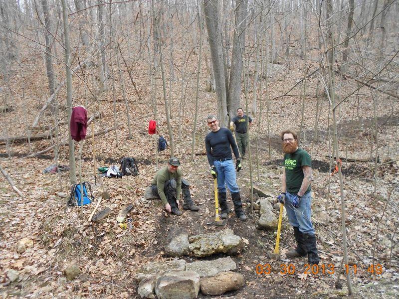 A group of five volunteers working together in a forested area, engaged in trail maintenance. They are using shovels and other tools to clear and improve a path made of rocks. Surrounding them are bare trees and fallen leaves, indicating early spring. Some backpacks and clothing items are visible in the background. Lost Valley mountain bike trail.