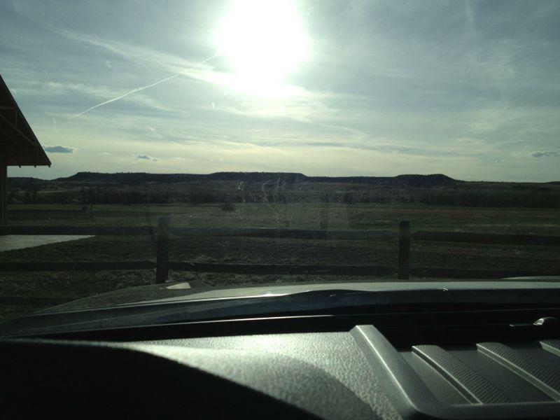 A scenic view of a vast landscape under a bright sky, with rolling hills in the background and a wooden fence in the foreground. The sunlight is shining brightly, creating a warm atmosphere, while clouds drift across the sky. The view is framed by the edge of a vehicle's dashboard, indicating the perspective from inside a car. Hidden Mesa mountain bike trail.