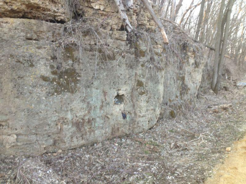 Rocky cliffside with exposed earth and vegetation, featuring a small hole in the rock face and bare trees in the background. The surrounding area is rugged and natural, with a dirt path along the base. Heritage Trail mountain bike trail.