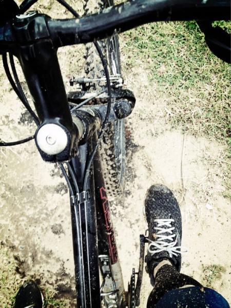 A close-up view of a mountain bike on a dirt path, with a muddy front wheel and a foot in sneakers resting on the pedal. The bike's handlebar and part of the frame are visible in the foreground, along with uneven ground and patches of grass. Cerro Gordo Trail mountain bike trail.