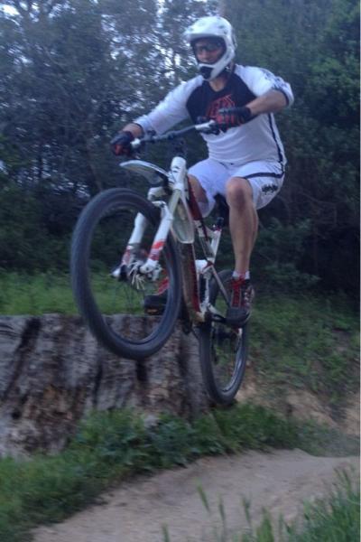 A cyclist in a white and black jersey and helmet is performing a jump on a mountain bike over a small hill or dirt mound, with greenery in the background. The bike's front wheel is off the ground as the rider is airborne, showcasing a dynamic action shot. China Camp mountain bike trail.