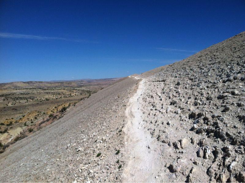 A rocky slope with a winding dirt path leading upward, surrounded by sparse vegetation and open land under a clear blue sky. The landscape features rolling hills and distant mountains. White Ridge Bike Trails mountain bike trail.