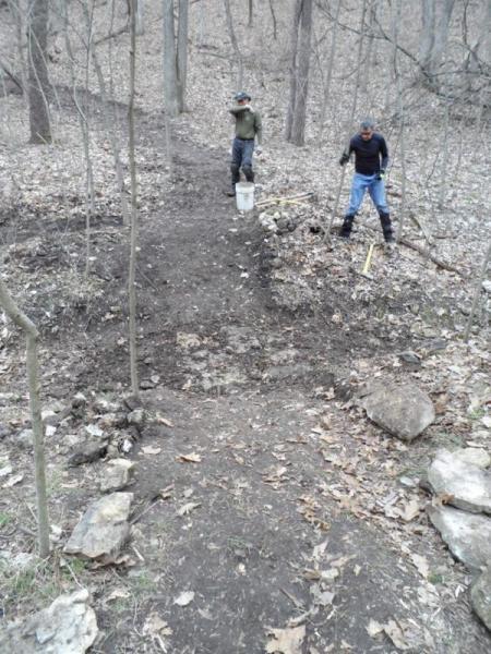 Two individuals are working on a hiking trail in a wooded area. One person is standing on a dirt path, holding a tool, while the other is near a bucket on the ground. The surrounding environment features bare trees and scattered fallen leaves, indicating a spring or late fall season. A few rocks are visible along the trail edge. Lost Valley mountain bike trail.