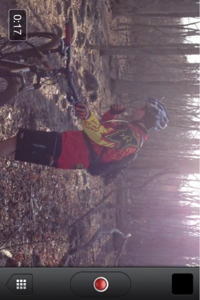 A cyclist wearing a red and yellow jersey, black shorts, and a helmet stands next to his mountain bike in a forested area. Sunlight filters through the trees, illuminating the ground covered in leaves and twigs. The cyclist appears to be adjusting something on the bike. Conway Robinson State Forest mountain bike trail.