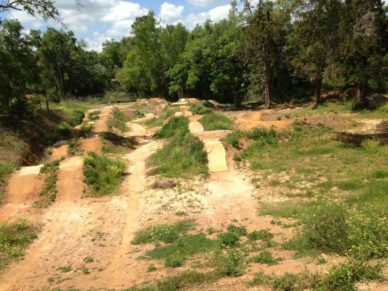 A dirt bike park featuring multiple mound jumps and trails surrounded by lush green trees and blue skies. The landscape shows slightly worn paths and patches of grass, indicating usage by cyclists. Tom Brown / Lafayette Heritage Park mountain bike trail.