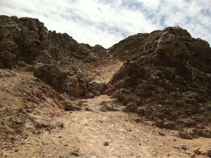 A rugged rocky pathway leading up through steep terrain, surrounded by mountains under a partly cloudy sky. The ground is dry and barren, with scattered rocks and minimal vegetation. White Ridge Bike Trails mountain bike trail.