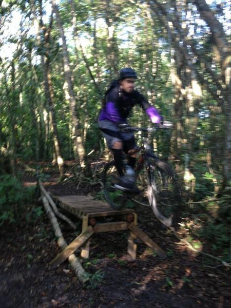 A mountain biker in a helmet and riding gear jumps off a wooden ramp in a forested area. The biker is mid-air with the bike lifted, surrounded by trees and greenery. The image is slightly blurred, capturing the motion and excitement of the jump. Tortuguero mountain bike trail.