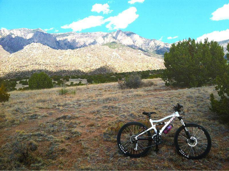 A mountain bike rests on grassy terrain with a backdrop of rugged mountains under a partly cloudy sky. The landscape features shrubs and sparse vegetation, creating a serene outdoor atmosphere ideal for cycling adventures. South Foothills mountain bike trail.