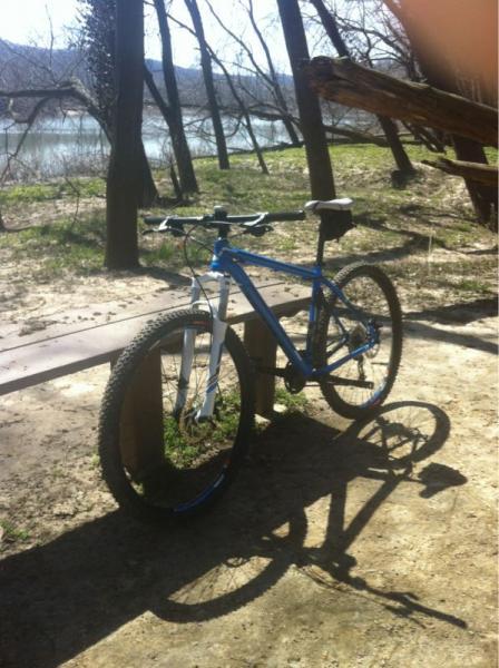 A blue mountain bike parked on a wooden bench near a river. The background features trees and a grassy area, with sunlight casting shadows on the ground. Castlewood State Park mountain bike trail.