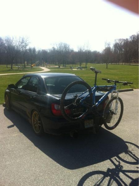 A black car parked in a sunny location with a bicycle attached to a bike rack on its rear. The background features a grassy area with bare trees and a path, suggesting a park setting. Castlewood State Park mountain bike trail.