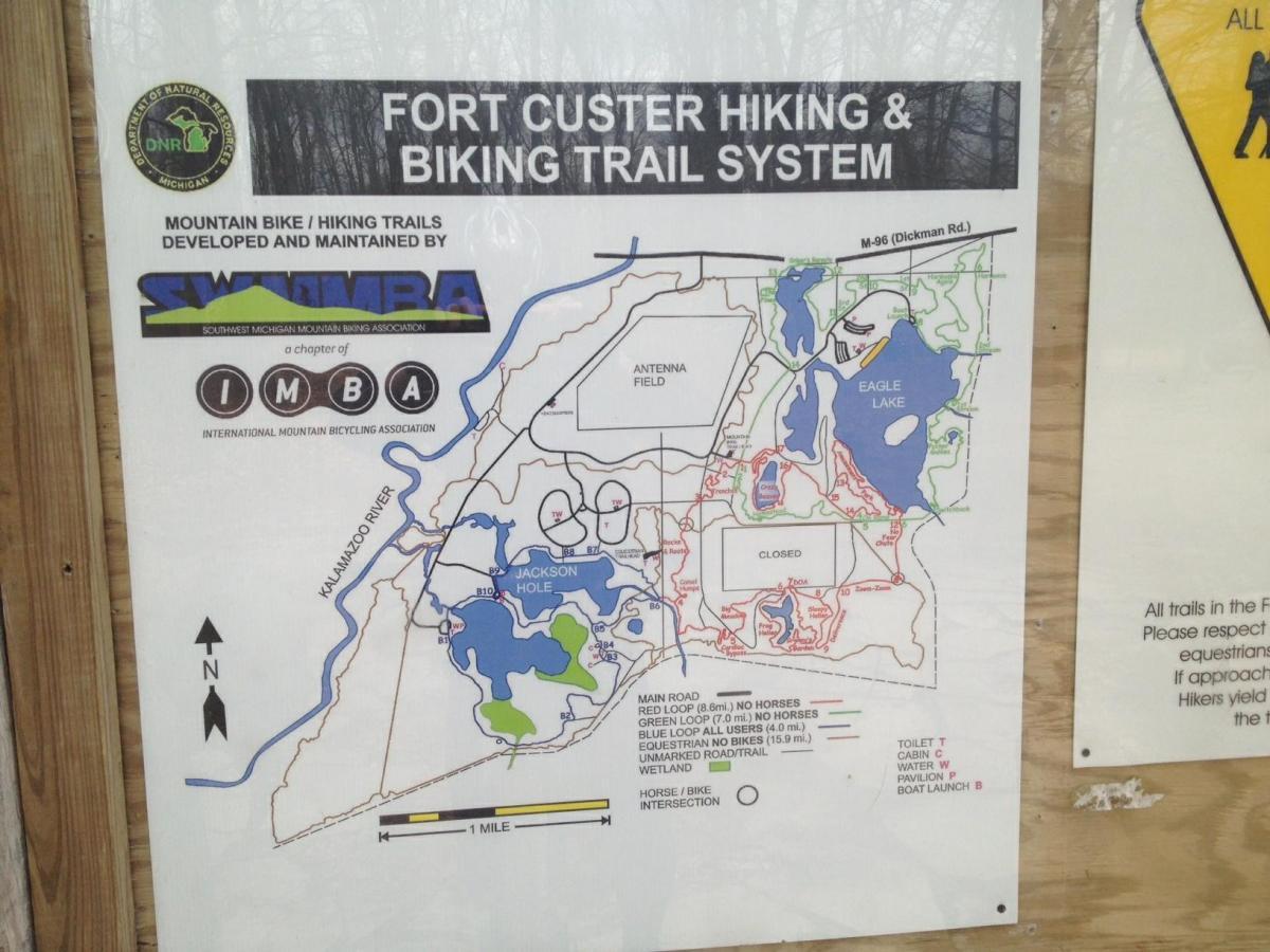 Map of the Fort Custer Hiking and Biking Trail System, showing various trails for mountain biking and hiking. Key features include trail names, loop distances, and trail usage restrictions. Additional information includes locations for restrooms, water access, cabin, and boat launch. The map is positioned against a wooden background in a forested area. Fort Custer Recreation Area mountain bike trail.