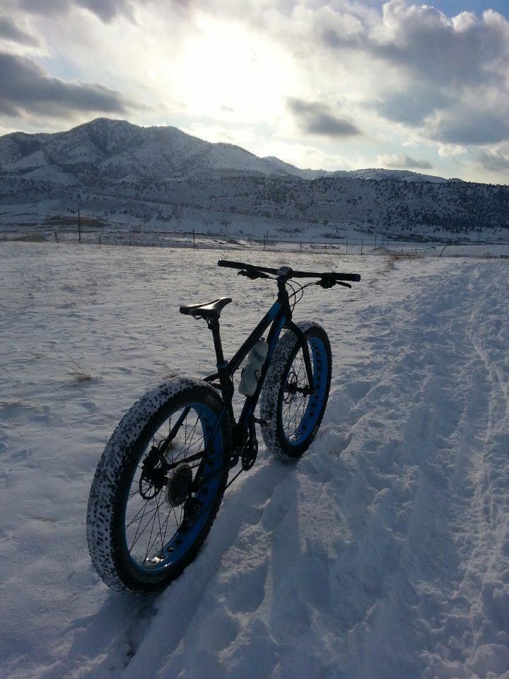 A fat bike resting on a snow-covered trail, with mountains in the background and a cloudy sky illuminated by the sun. Green Mountain mountain bike trail.