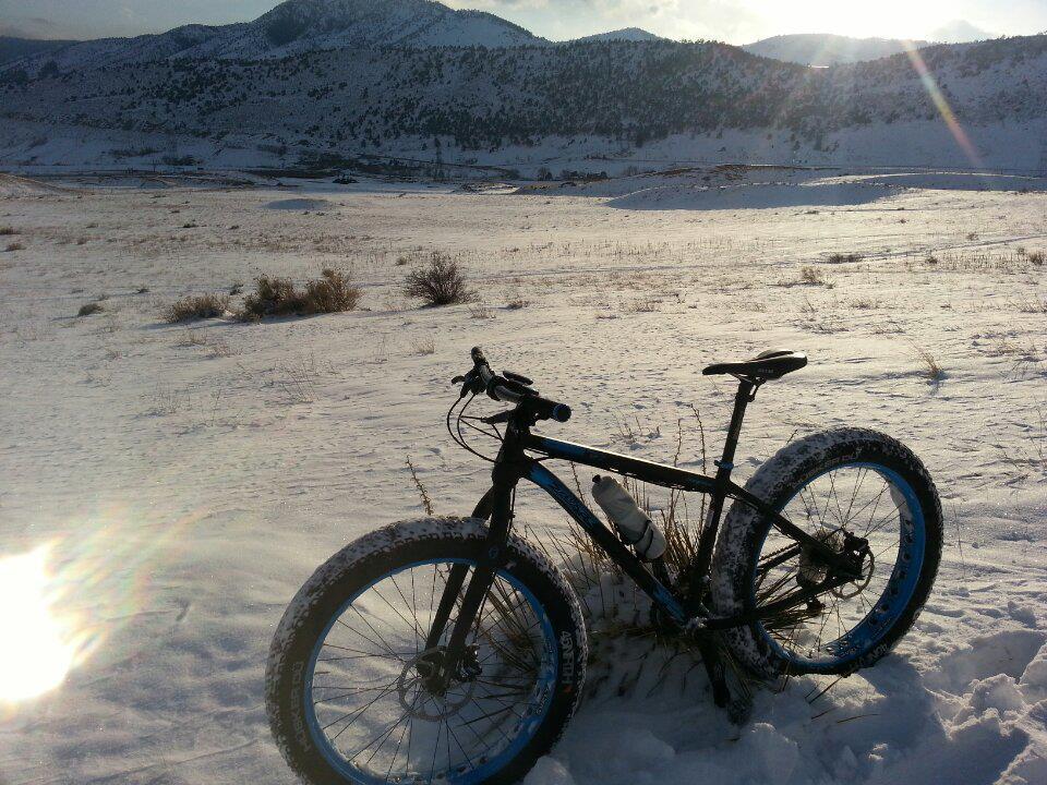 A black and blue fat bike is parked on a snowy landscape, with mountains visible in the background. The ground is covered in white snow, and the sunlight creates a shimmering effect on the surface. The bike's large tires have a layer of snow on them, indicating recent use. Green Mountain mountain bike trail.