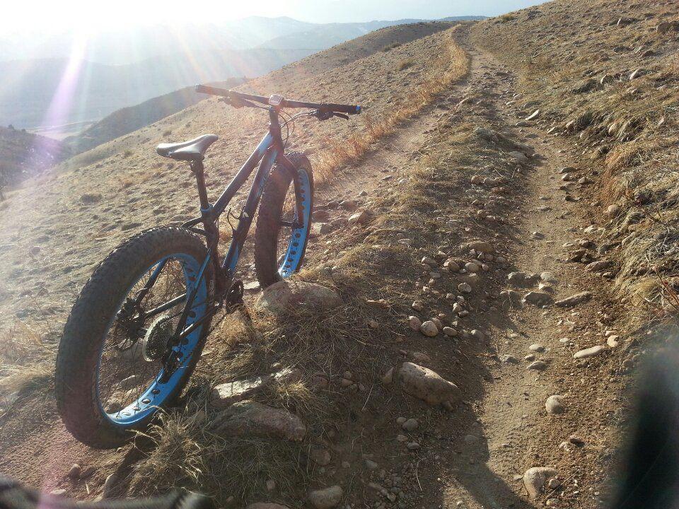 A fat tire bike parked on a rocky trail in a mountainous area, with sunlight streaming through the lens creating a bright, scenic view of the surrounding landscape. The trail is made of dirt and scattered stones, indicating a rugged terrain perfect for biking adventures. Green Mountain mountain bike trail.