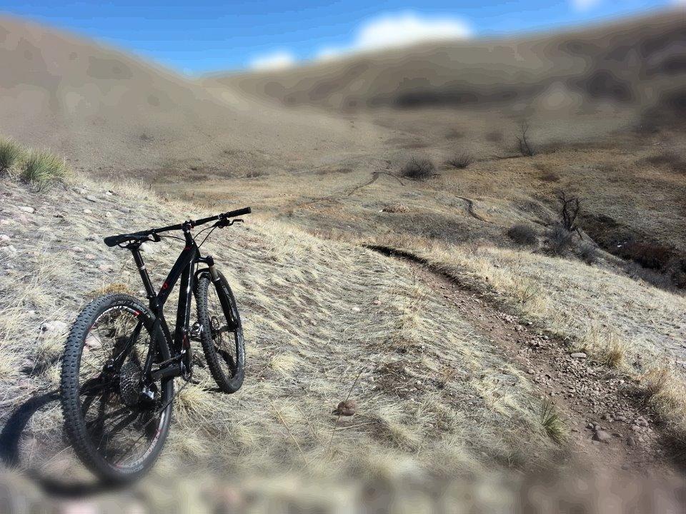 A mountain bike leaning against a grassy slope, with a dirt trail winding through a hilly landscape under a blue sky with scattered clouds. Green Mountain mountain bike trail.