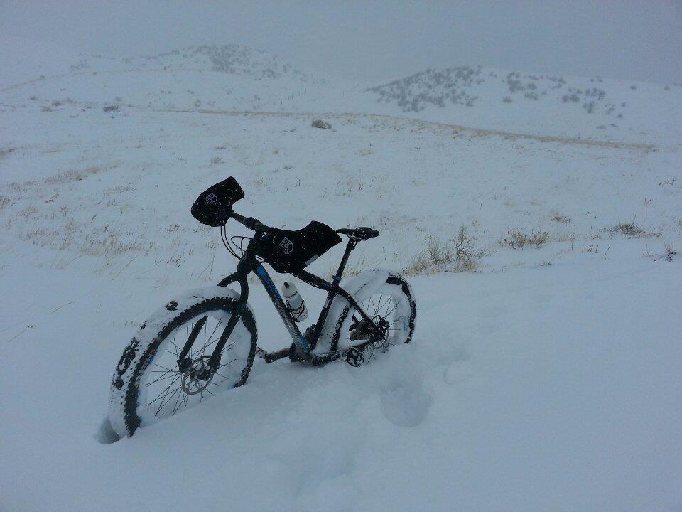 A mountain bike partially buried in snow on a wintery landscape, with snow-covered hills in the background and a cloudy sky above. Green Mountain mountain bike trail.