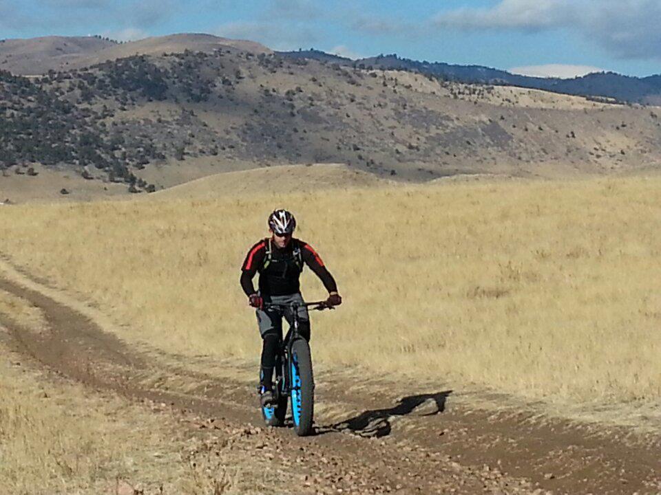 A cyclist riding a fat bike on a dirt path through a grassy landscape, with rolling hills and a blue sky in the background. The cyclist is wearing a black and red long-sleeve shirt, a helmet, and padded shorts. Green Mountain mountain bike trail.