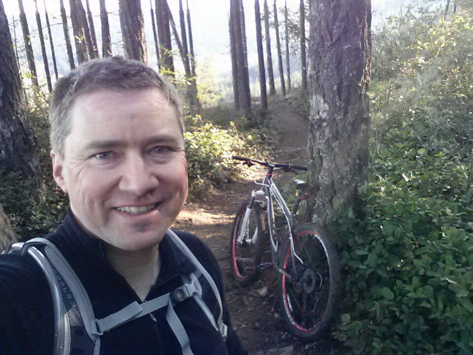 A person smiling for a selfie while standing on a wooded trail with tall trees in the background. A mountain bike is leaning against a tree nearby. The scene is bright and outdoorsy, suggesting an enjoyable day of biking and exploring nature. Green Mountain Trails mountain bike trail.