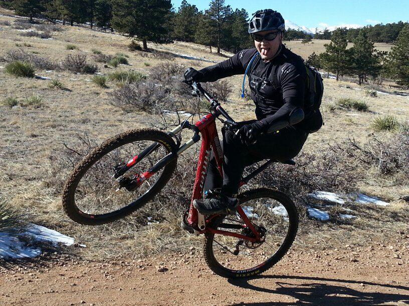 A person wearing a helmet and black cycling gear performs a wheelie on a red mountain bike on a dirt trail surrounded by grassy and rocky terrain, with trees and mountains in the background. The cyclist is sticking out their tongue, expressing excitement. Hall Ranch mountain bike trail.
