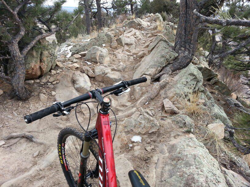 Mountain bike handlebars in the foreground, overlooking a rocky, uneven trail surrounded by trees and sparse vegetation. The trail is rugged with exposed roots and loose stones, indicating a challenging biking route. Red Rocks / Dakota Ridge mountain bike trail.