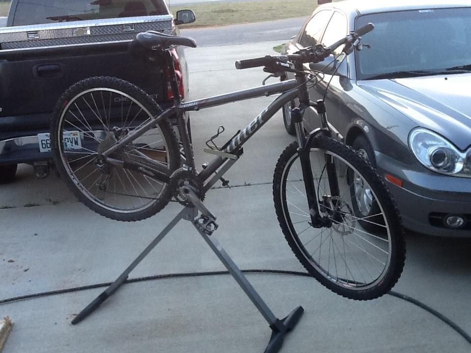 Niner EMD 9: A mountain bike is mounted on a bike repair stand, with its front wheel elevated. In the background, there are two parked vehicles: a black pickup truck and a gray sedan. The scene is set outdoors on a concrete driveway.