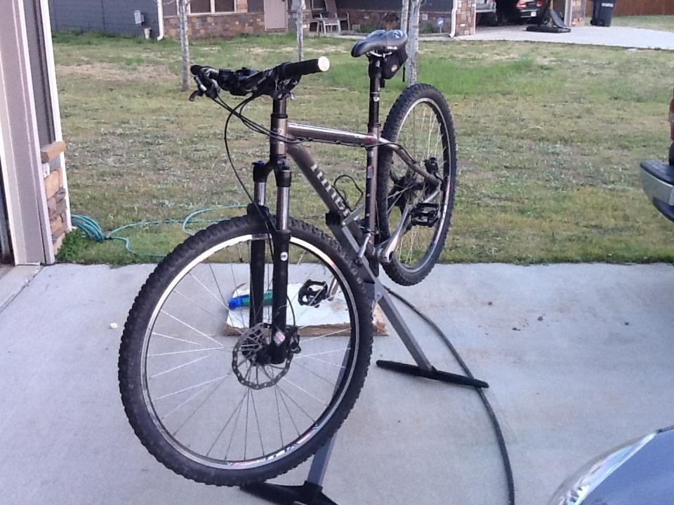 Niner EMD 9: A mountain bike on a repair stand in a driveway, with a grassy area in the background. The bike has a gray frame, thick tires, and front suspension. The setting is a residential area, with a garage visible in the background.